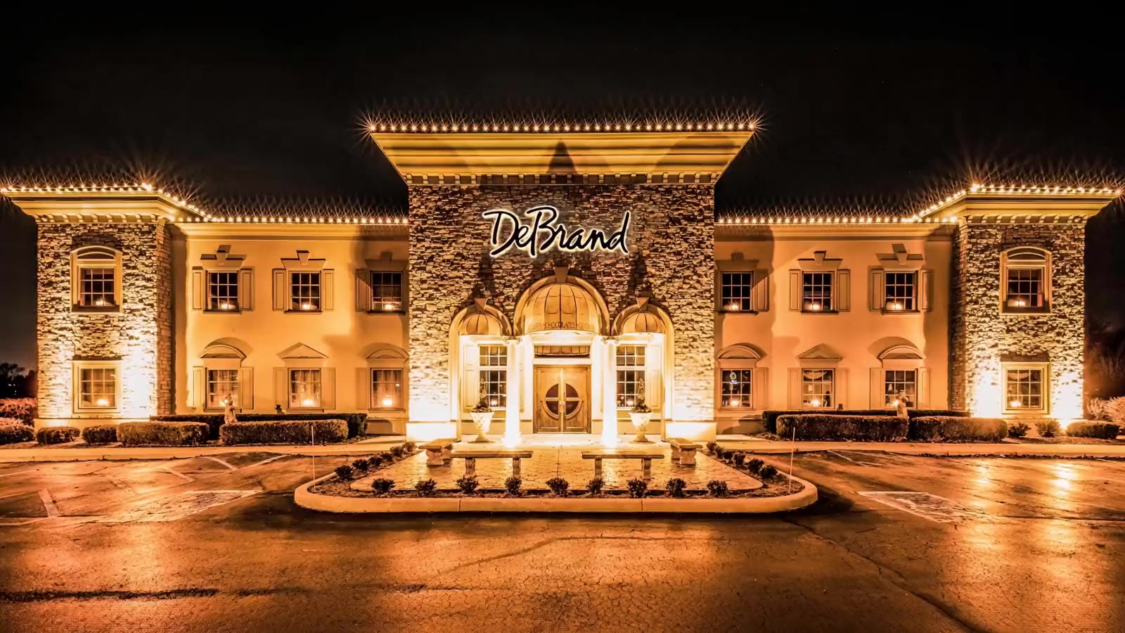 DeBrand's Auburn Road building at night: a stone and cream facade under warm light, string lights along every roofline, the script DeBrand sign above a columned entrance