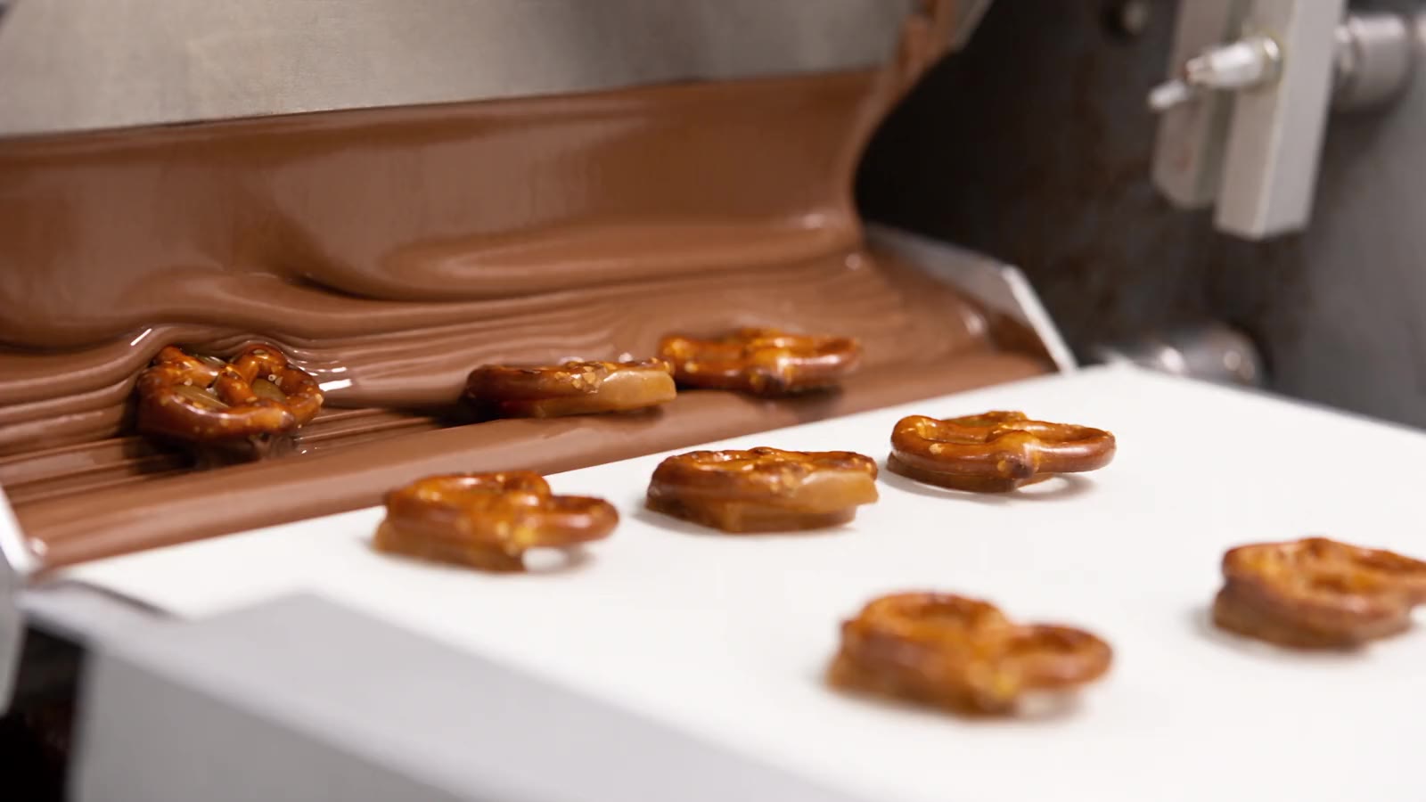 A caramel pretzel passing under a falling stream of milk chocolate on a white conveyor belt and emerging coated, with more bare pretzels queued behind it