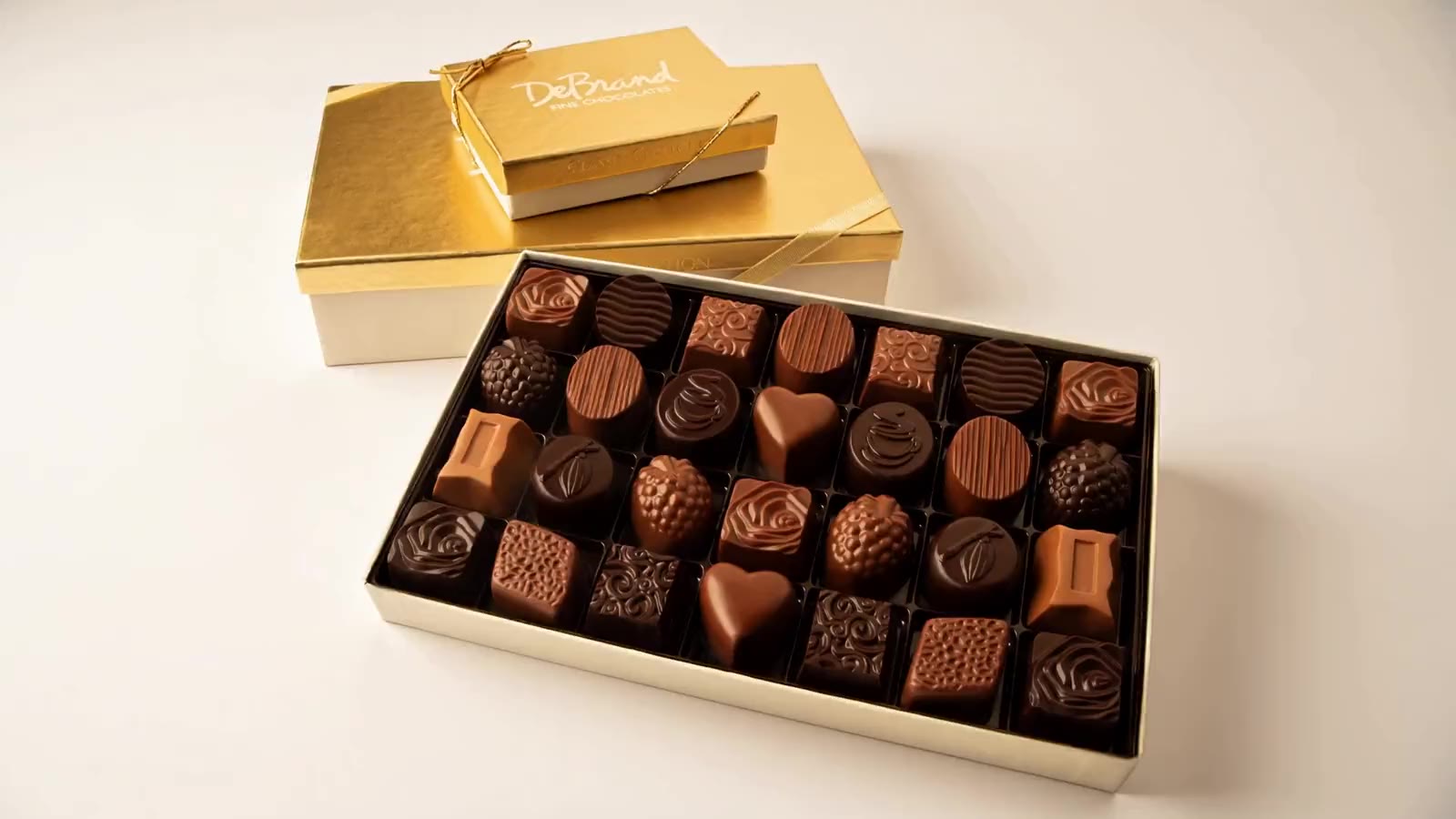 A gold-lidded cream box of assorted DeBrand chocolates, tied with a gold cord, beside a smaller gold box